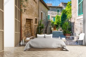 Wizualizacja fototapety przedstawiająca spokojną uliczkę ozdobioną kwiatami i zieloną roślinnością, oraz schody do starej, włoskiej kamienicy. Fototapeta do pokoju dziennego, sypialni, salonu, przedpokoju i jadalni, biura lub gabinetu, optycznie powiększająca pomieszczenie.